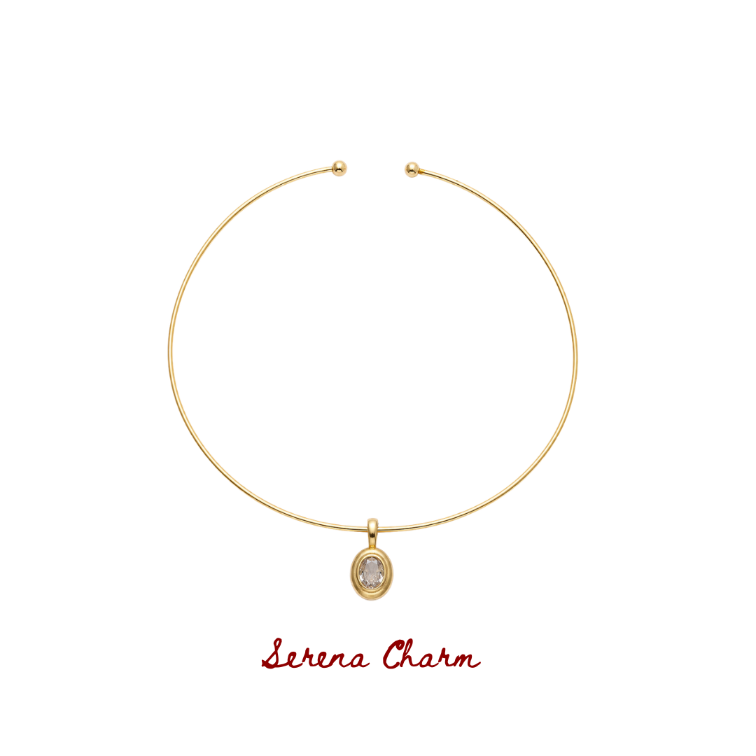 A gold wire bangle necklace with a small oval charm featuring a clear stone, displayed on a white background with the text 'Serena Charm' written in red below.